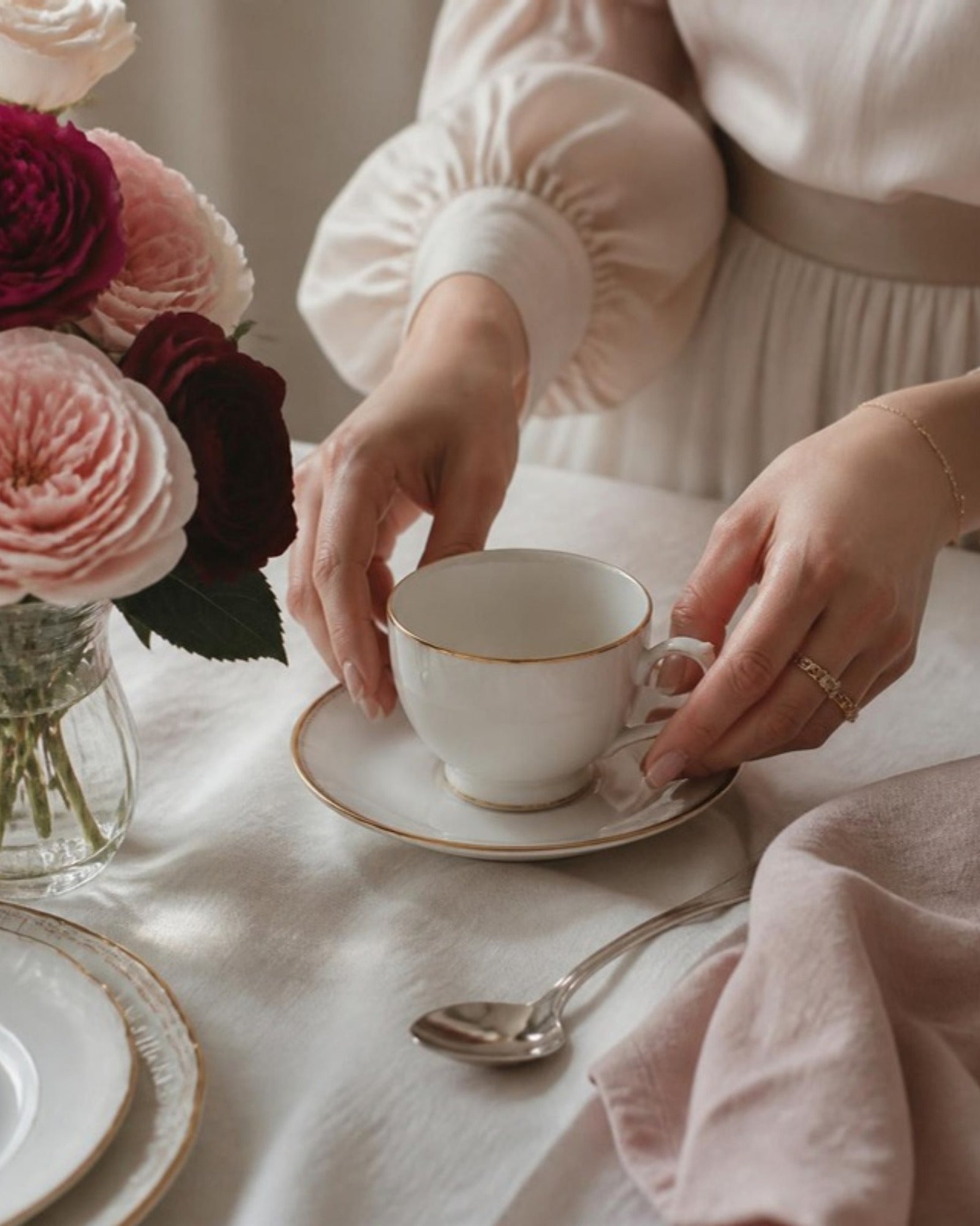 Hands setting a gold rimmed bone china teacup on a linen tablecloth beside garden roses